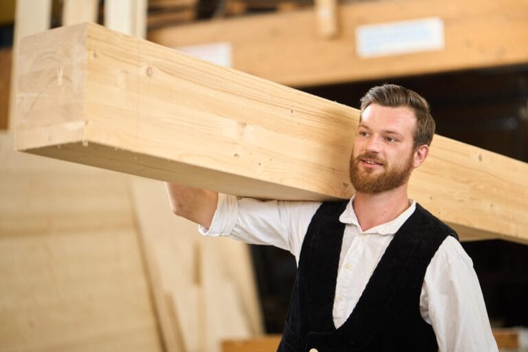 Zimmerer trägt massiven Holzträger – Mitarbeiterfotos, Industriefotografie und Handwerksbetrieb, Rhein‑Neckar‑Region.
