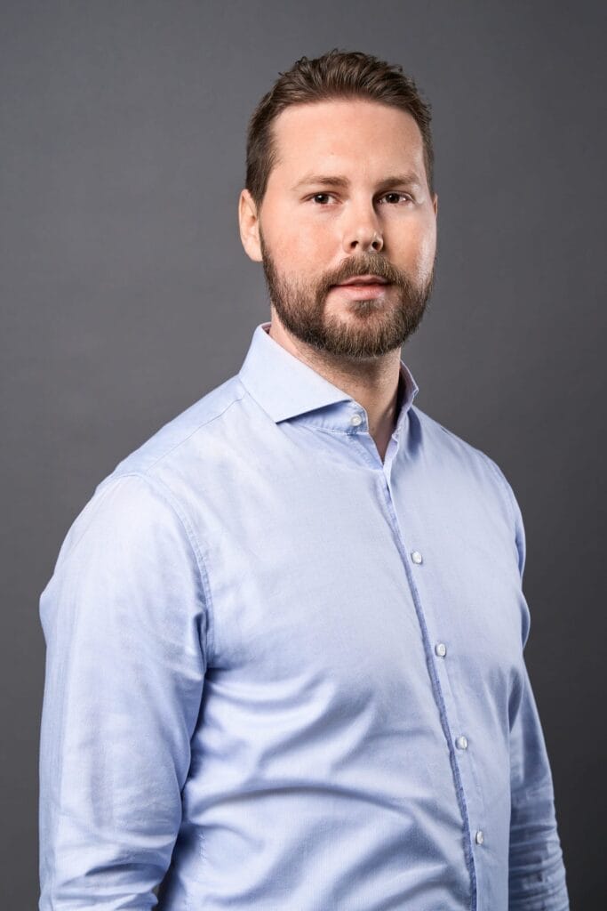 Ein Mann mit kurzen braunen Haaren und Bart, der ein hellblaues Button-up-Hemd trägt, steht vor einem schlichten grauen Hintergrund. Dieses Bild fängt den für die Mitarbeiterfotografie Siemens typischen professionellen Stil ein, da er mit einem neutralen Ausdruck leicht zur Seite blickt.