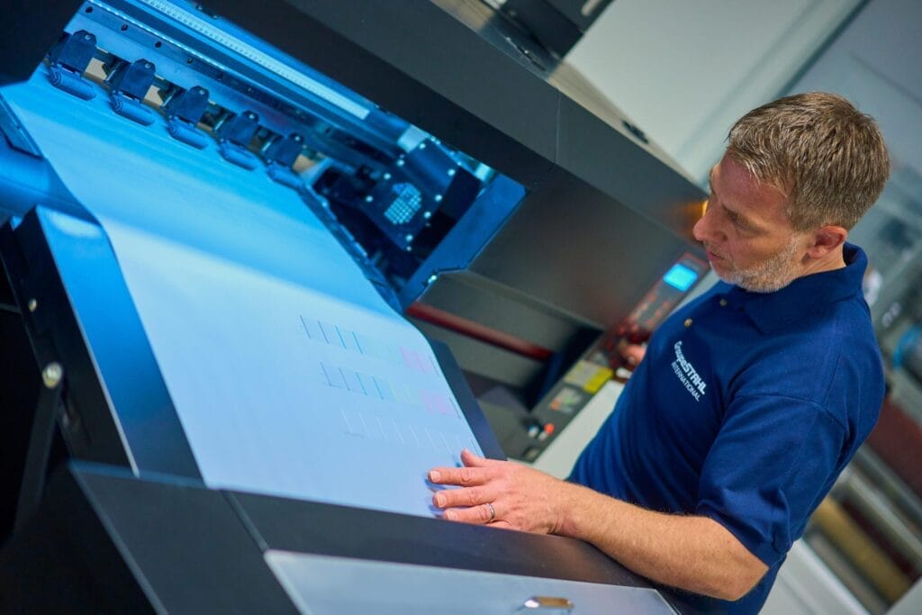 Ein Mann in einem blauen Polohemd prüft einen großen Papierbogen, der aus einem Industriedrucker bei Stahls' Europe kommt, der professionelle Businessfotografie in einer modernen saarländischen Druckerei zeigt.