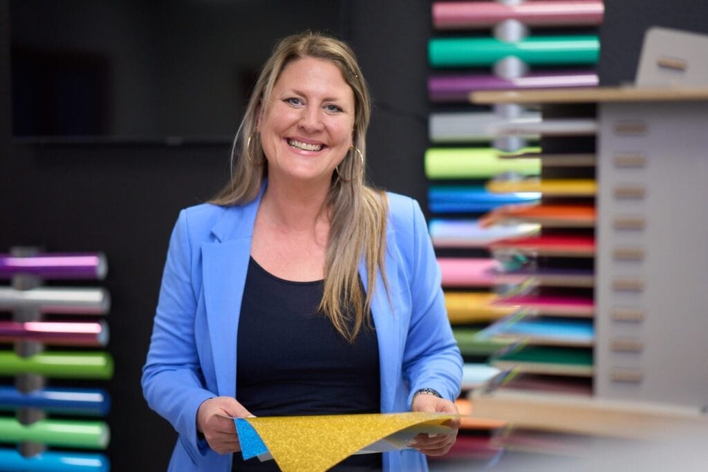 Eine lächelnde Frau in einem blauen Blazer hält ein Blatt gelbes Glitzerpapier in der Hand. Hinter ihr sind bunte Papierrollen in Regalen vor einem dunklen Hintergrund ausgestellt - ein Beispiel für lebendige Businessfotografie im Saarland.
