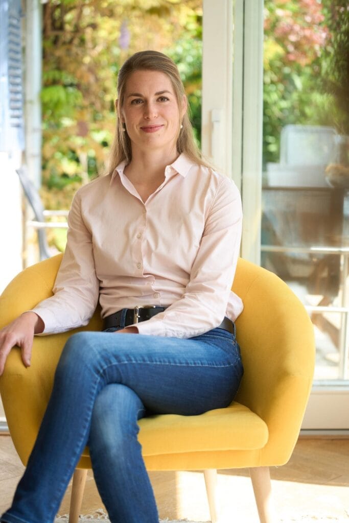 Eine Frau mit langen blonden Haaren, die ein hellrosa Hemd und eine blaue Jeans trägt, sitzt im Schneidersitz auf einem gelben Stuhl in einem hellen Raum mit großen Fenstern und viel Grün im Freien - eine ideale Umgebung für Personal Branding oder Businessfotografie.