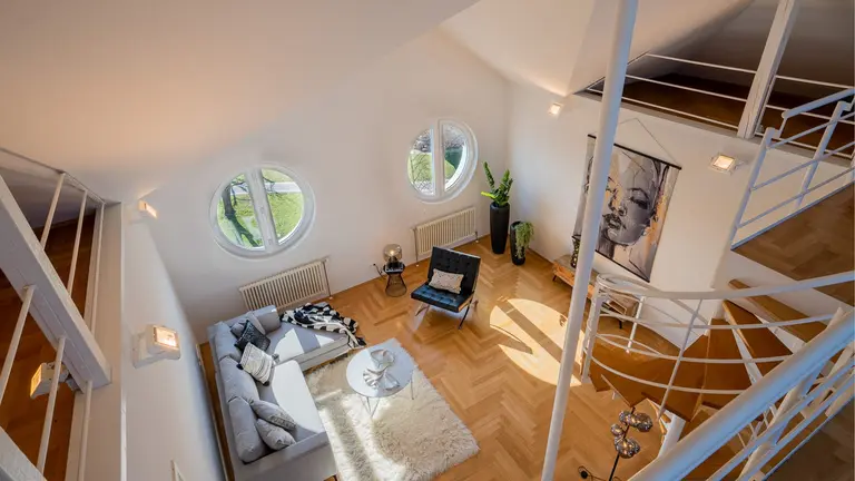 Modernes Wohnzimmer mit hellem Holzboden, grauem Sektionssofa, schwarzem Sessel, runden Fenstern und einer Wendeltreppe - aufgenommen in einem hellen, luftigen Raum in Ladenburg. Perfekte Inspiration für Businessfotografie oder Filmsetdesign.