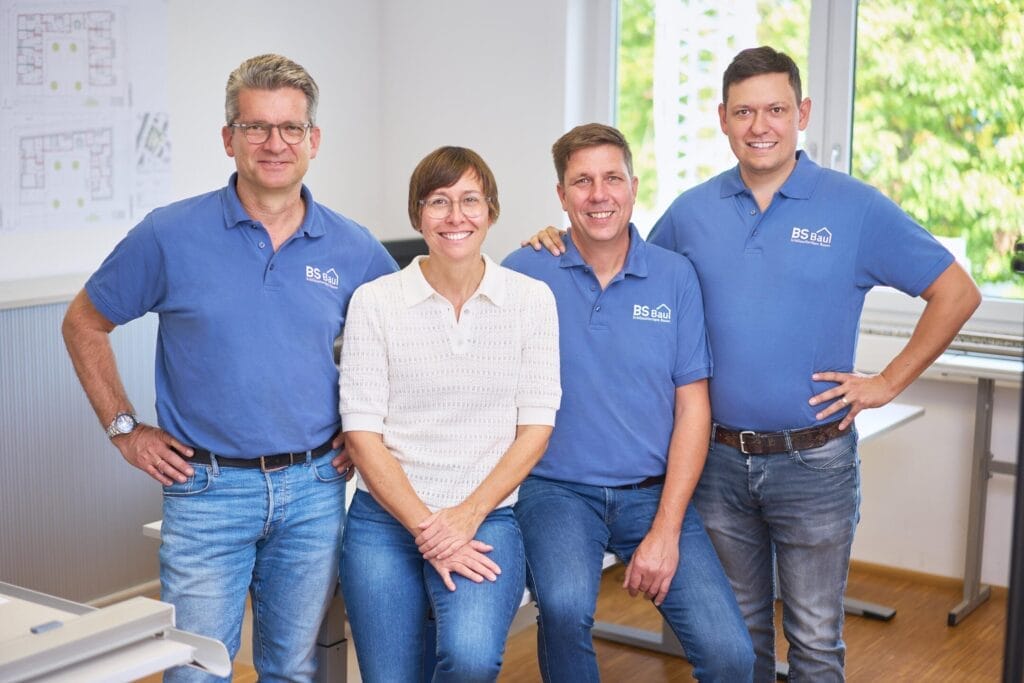 Vier Personen posieren und lächeln in einem hellen Büro, das Sauterbau repräsentiert. Drei Männer in passenden blauen Poloshirts stehen um eine Frau in einem weißen Top, die an einem Schreibtisch sitzt. Die Unternehmensfotografie hebt Pläne und Grünflächen hervor, die durch das Fenster zu sehen sind.