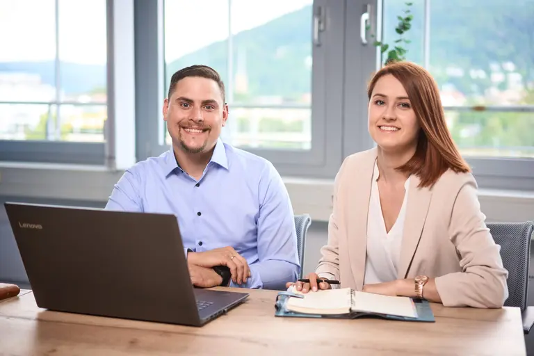 Zwei Personen sitzen mit Laptop und Notebook an einem Schreibtisch in einem Büro und lächeln in die Kamera. Große Fenster hinter ihnen bieten einen malerischen Blick ins Grüne, der den Geist der Brandingfotografie perfekt einfängt und Sie dazu einlädt, in Glück zu investieren.