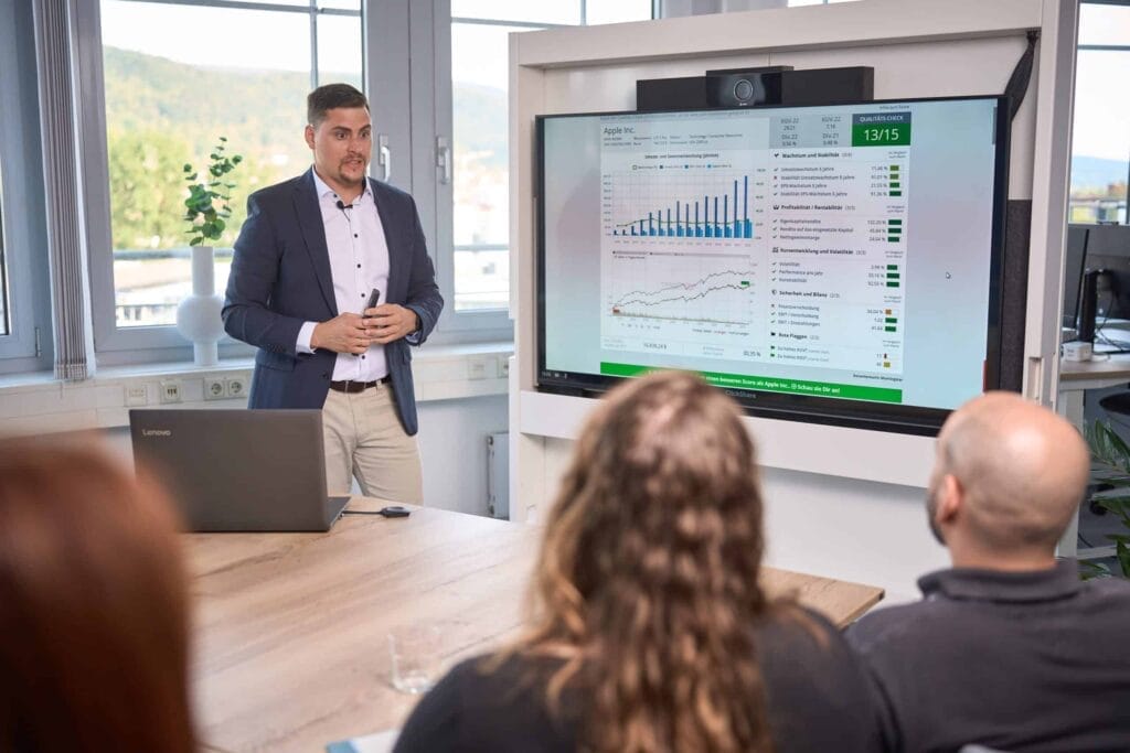 Ein Mann im Business-Outfit präsentiert drei sitzenden Personen in einem modernen Besprechungsraum mit großen Fenstern Datendiagramme auf einem großen Bildschirm, um die Wirkung von Brandingfotografie zu zeigen und zu erläutern, warum Unternehmen in Glück investieren sollten.