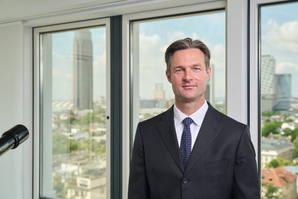 Ein Mann in dunklem Anzug und Krawatte steht in einem Büro mit großen Fenstern und Blick auf die Stadt - perfekt für Businessportraits Frankfurt, die die Präsenz einer Führungskraft vor der Kulisse hoher Gebäude und eines blauen Himmels einfangen.