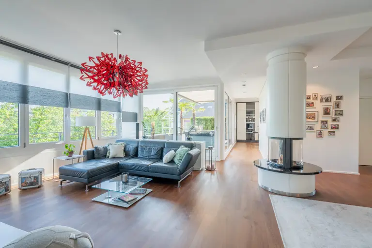Modernes Penthouse-Wohnzimmer mit großen Fenstern, einem schwarzen Sektionssofa, einem Couchtisch aus Glas, einem roten Kronleuchter und einem runden Kamin. Helle Wände und Holzböden schaffen eine helle, luftige Atmosphäre mit viel Grün - ideal für Immobilienfotografie in Heidelberg.