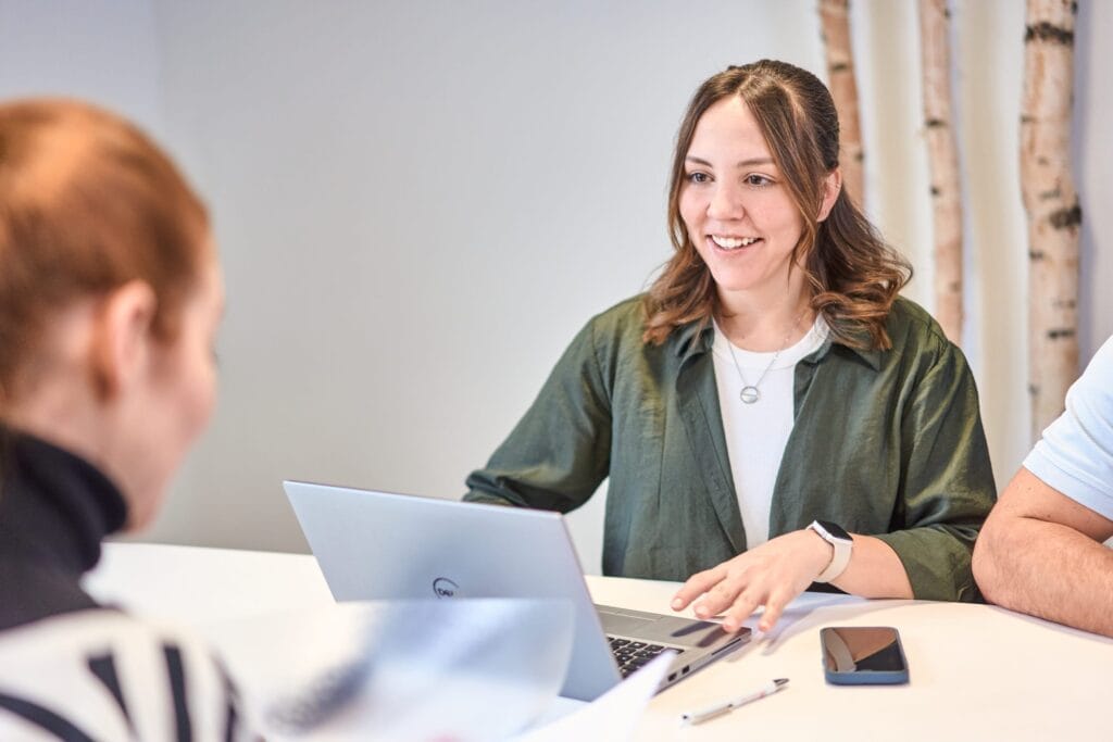 Eine Frau mit braunen Haaren und grünem Hemd sitzt mit Laptop, Smartphone und Stift an einem Tisch, lächelt und unterhält sich mit einer anderen Person - und fängt damit die Essenz der Brandingfotografie für die Bürstadt Furniture GmbH ein.