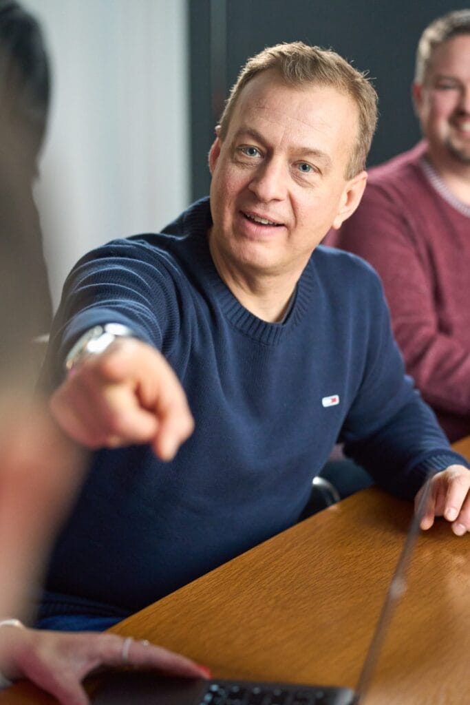 Ein Mann in einem marineblauen Pullover sitzt an einem Tisch, lächelt und deutet auf jemanden im Off-Kamera-Bereich. Eine andere Person in einem kastanienbraunen Pullover sitzt neben ihm, beide genießen ein zwangloses Treffen - ideale Mitarbeiterfotos für den Bedarf von vobapay an authentischen Brandingfotos.