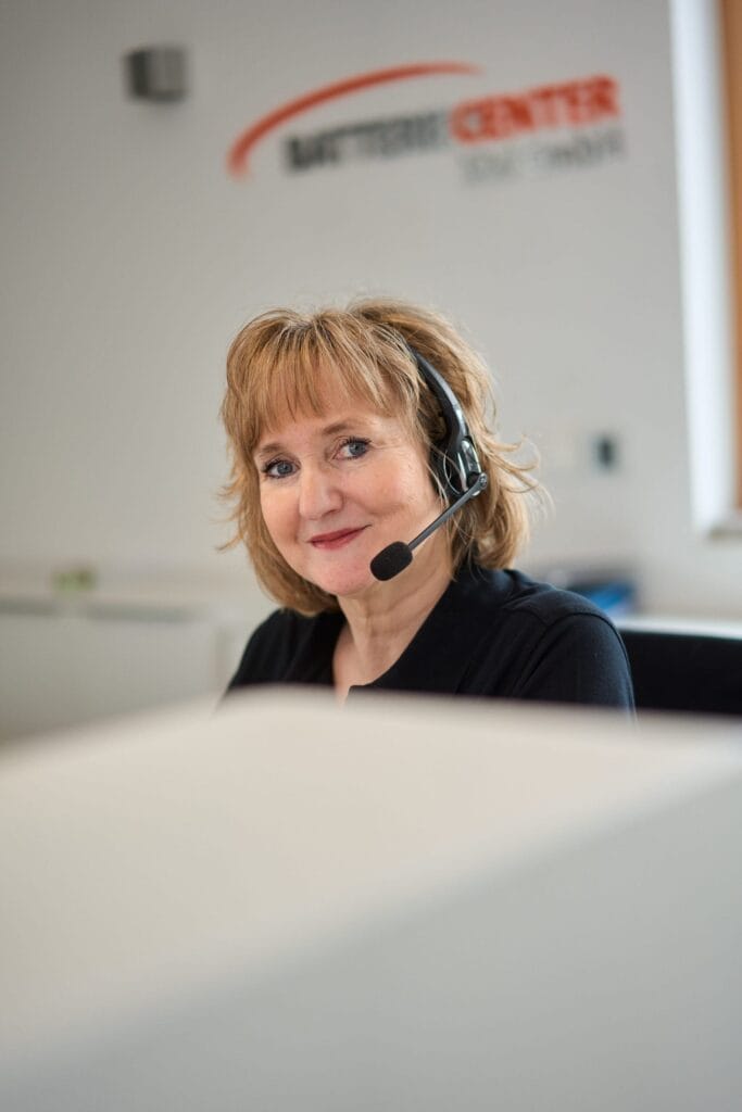 Eine Frau mit Headset sitzt lächelnd an ihrem Schreibtisch im Büro des Batteriecenter Süd. Im Hintergrund ist ein verschwommenes Logo an der Wand zu sehen, das authentische Mitarbeiterfotografie und subtile Brandingfotografie einfängt.