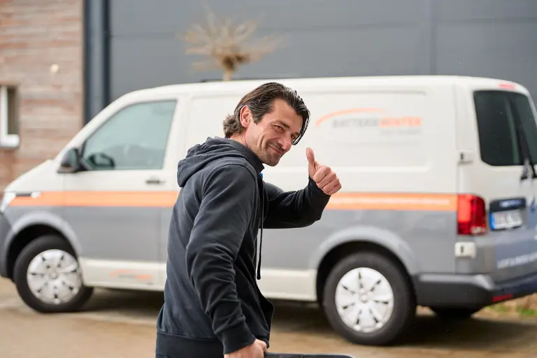 Ein Mann in einem schwarzen Kapuzenpulli lächelt und zeigt den Daumen nach oben, während er vor einem weißen Lieferwagen mit orangefarbener Beschriftung steht - perfekt für die Mitarbeiterfotografie im Batteriecenter Süd. Im Hintergrund sind ein Gebäude und ein kleiner Baum zu sehen.