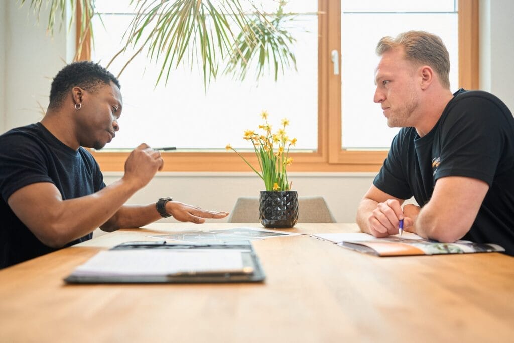 Zwei Männer sitzen sich an einem Holztisch gegenüber und diskutieren für die Mitarbeiterfotografie im Batteriecenter Süd. Der eine gestikuliert mit einem Stift, der andere hört zu, zwischen ihnen liegen Papiere und eine Topfpflanze, während das Sonnenlicht durch das Fenster dahinter fällt.