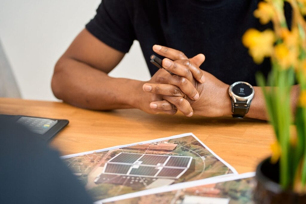 Eine Person, die eine Armbanduhr trägt, sitzt mit gefalteten Händen an einem Tisch und hält einen Stift in der Hand. Auf dem Tisch liegen Architekturpläne - perfekt für Mitarbeiterfotografie oder Brandingfotografie - während im Vordergrund eine verschwommene gelbe Blume erscheint.