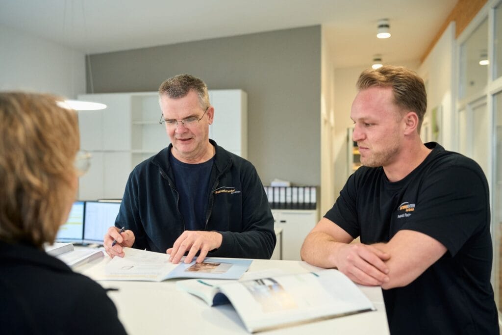 Zwei Männer sitzen an einem weißen Schreibtisch, prüfen Dokumente und unterhalten sich mit einer Frau, die mit dem Rücken zur Kamera steht, in einer modernen Büroumgebung im Batteriecenter Süd - ein Beispiel für professionelle Mitarbeiterfotografie.