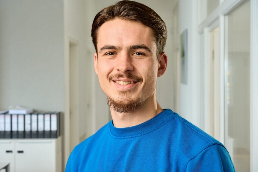Ein junger Mann mit kurzen braunen Haaren und gestutztem Bart steht lächelnd in einem Innenraum und verkörpert die professionelle mitarbeiterfotografie für Batteriecenter Süd. Er trägt ein hellblaues Hemd, im Hintergrund sind Regale und weiße Wände zu sehen.