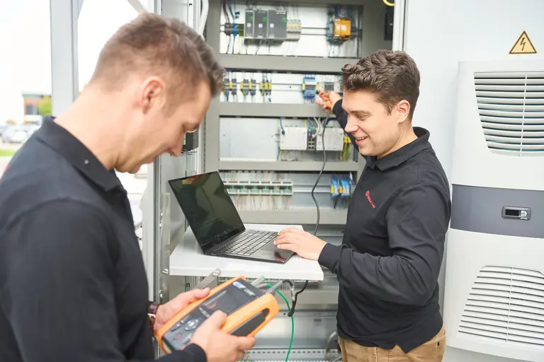 Zwei Techniker der Edisol GmbH arbeiten vor einem offenen Schaltschrank mit Laptop und Prüfgerät an elektrischen Geräten. Diese Industriefotografie-Szene wurde am Standort Ravensburg aufgenommen.