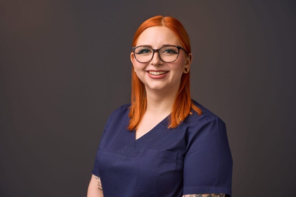 Eine Frau mit glattem roten Haar, Brille und Nasenring lächelt in die Kamera. Sie trägt einen blauen Arztkittel und steht vor einem schlichten dunklen Hintergrund - perfekt für die Mitarbeiterfotografie in einer modernen Arztpraxis.