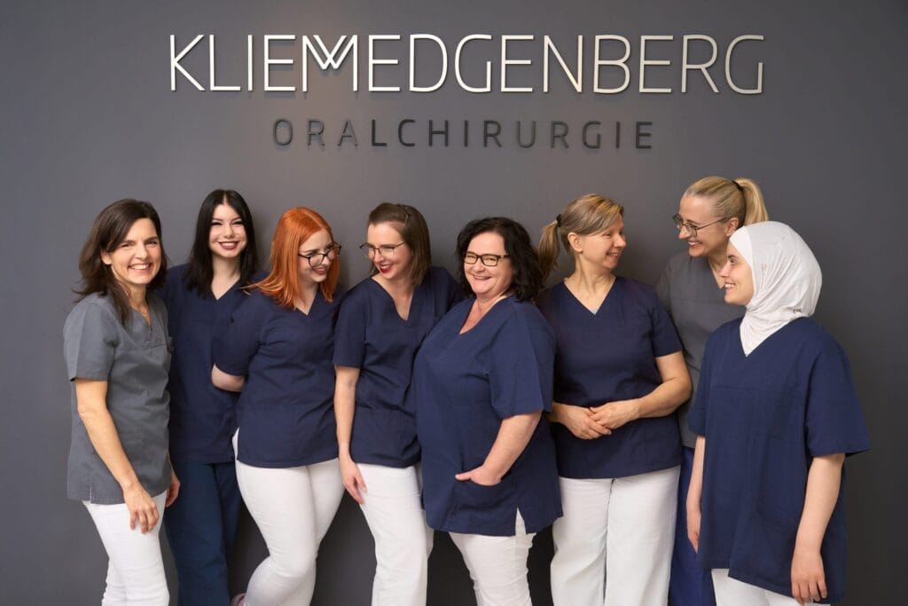 Acht Frauen in passenden Arztkitteln stehen lächelnd und lachend zusammen vor einem Wandschild mit der Aufschrift Kliem Edgenberg Oralchirurgie - ein perfektes Beispiel für Mitarbeiterfotografie in der Arztpraxis. Der Hintergrund ist einfarbig dunkelgrau.