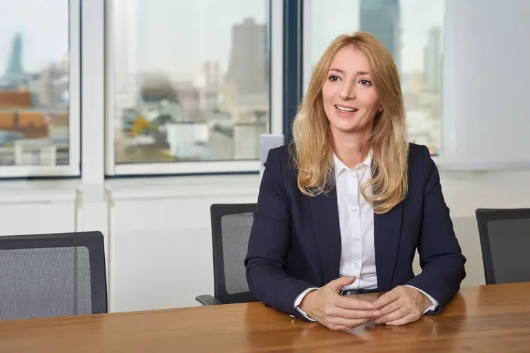 Eine Frau mit langen blonden Haaren, marineblauem Blazer und weißem Hemd sitzt lächelnd an einem Konferenztisch. Hinter ihr ist durch große Fenster eine Stadtlandschaft zu sehen - perfekt für Businessportraits Frankfurt.